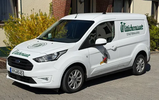 Weißer Lieferwagen mit Firmenlogo und Grafiken auf der Seite, geparkt auf einer Einfahrt vor einem Wohnhaus bei Tageslicht