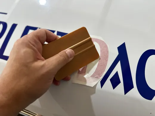 Eine Hand bringt mit einer Rakel ANKERHELD-Folienbuchstaben auf einer glatten Oberfläche an. © Kellermanns