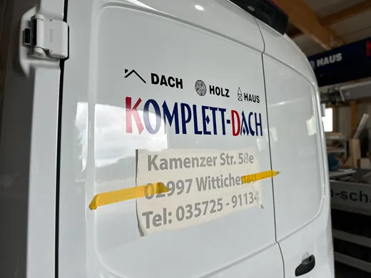 Seitenansicht eines weißen Firmenwagens mit „KOMPLETT-DACH“ Beschriftung, Adresse und Telefonnummer, inklusive ANKERHELD Logo. © Kellermanns