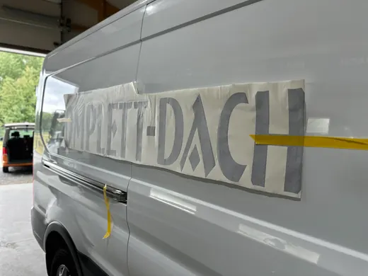 Ein weißer Transporter mit einer angeklebten Folie, auf der „KOMPLETT-DACH“ steht, daneben das Markenlogo