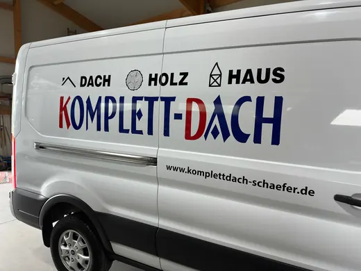 Ein weißer Transporter mit der Aufschrift „KOMPLETT-DACH“, Dach-Holz-Haus und der Marke ANKERHELD, daneben eine Webadresse. © Kellermanns