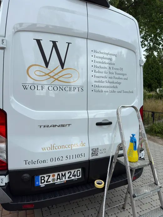 Rückseite eines weißen Lieferwagens mit Werbeaufschrift „WOLF CONCEPTS“, Leiter und Reinigungsmittel stehen daneben, Bäume im Hintergrund