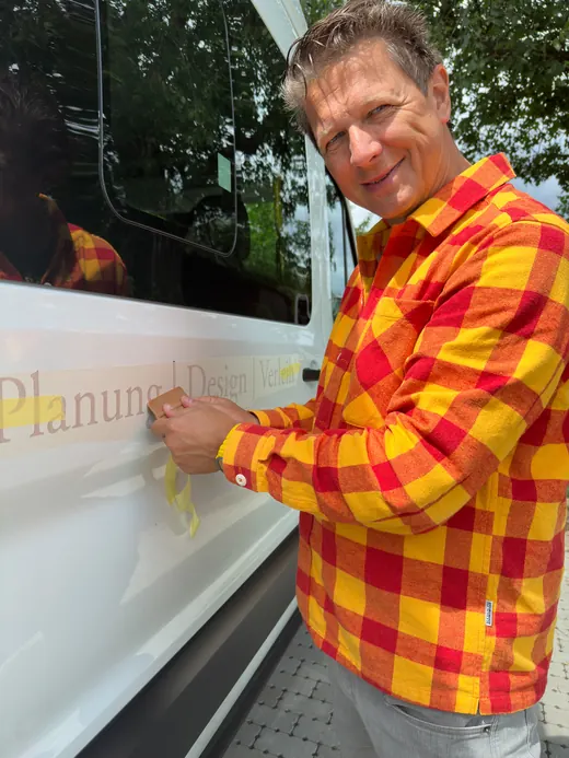 Ein Mann in kariertem Hemd klebt Schrift auf die Seite eines weißen Fahrzeugs und lächelt dabei in die Kamera