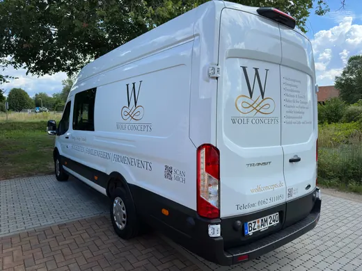 Weißer Lieferwagen mit schwarzem Logo und Schriftzug „W Bestattungen“ auf der Seite, steht auf gepflastertem Parkplatz unter Bäumen