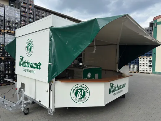 Weißer Bierwagen mit grünem Dach, ANKERHELD Logo und Aufschrift, steht auf einem freien Platz, Hochhäuser im Hintergrund, bereit für den Verkauf. © Kellermanns