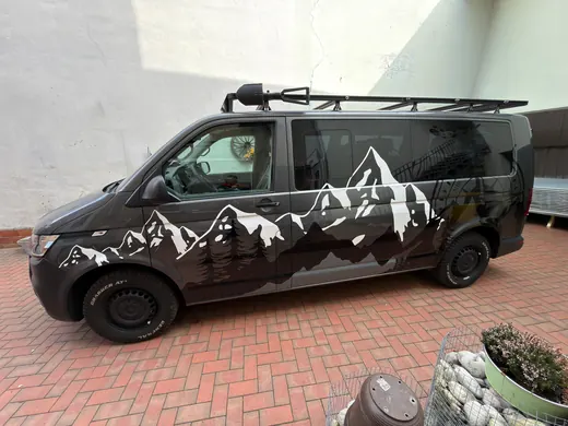 Schwarzer Van mit weißem Bergmotiv auf der Seite, auf einem gepflasterten Hof geparkt, Dachgepäckträger und einige Steine im Vordergrund