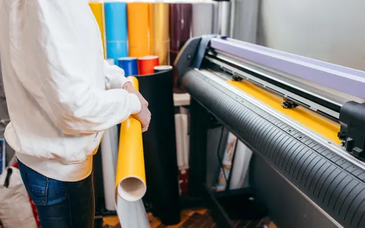 Person hält gelbe Folienrolle neben Schneideplotter, verschiedene bunte Folienrollen von ANKERHELD im Hintergrund. © Kellermanns