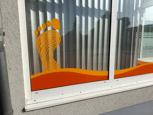 Orangefarbener Fußabdruck und Wellenmuster als Fensteraufkleber auf einer Glasscheibe, im Hintergrund reflektieren vertikale Lamellen und Gebäude