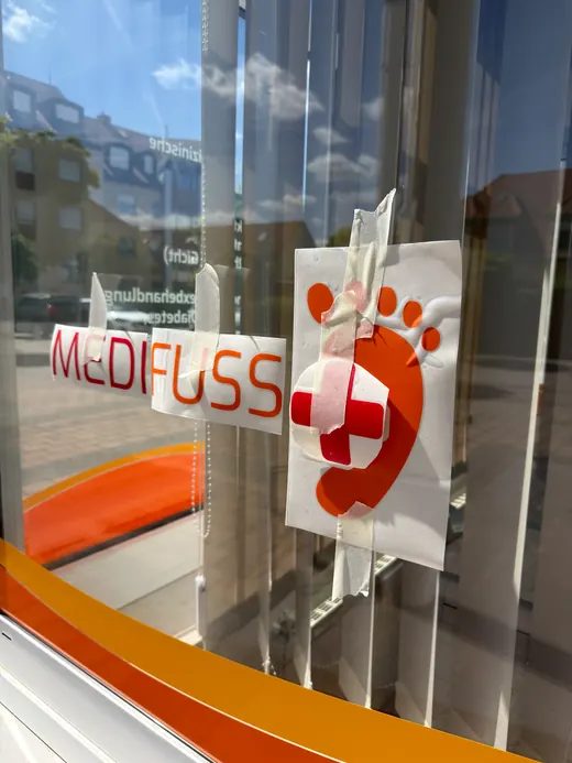 Schaufenster mit Schriftzug „MEDIFUSS“ und einem Plakat mit Fußsymbol, Kreuz und Pflaster, Spiegelungen von Gebäuden und Himmel im Glas