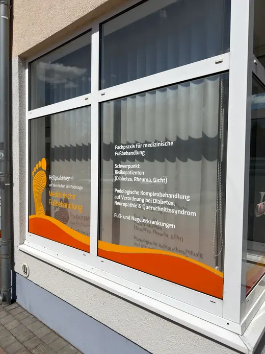 Schaufenster mit weißem Text und orangefarbener Grafik, Informationen über eine Sozialberatungsstelle, reflektierende Scheibe mit Vorhängen im Hintergrund