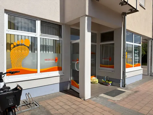 Außenansicht eines modernen Gebäudeeingangs mit großen Fenstern, bunten Grafiken und Pflanzen vor der Tür. Ein Fahrradständer steht an der Wand