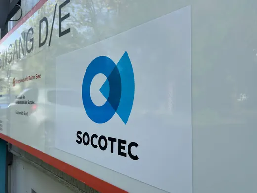 SOCOTEC-Logo auf einem weißen Schild mit blauem Symbol, daneben die Marke ANKERHELD, spiegelt sich leicht in einer Glasscheibe. © Kellermanns