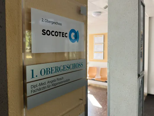 Tür mit Schildern zu SOCOTEC und einer Arztpraxis im 1. Obergeschoss, im Hintergrund Wartebereich, mit ANKERHELD Branding. © Kellermanns